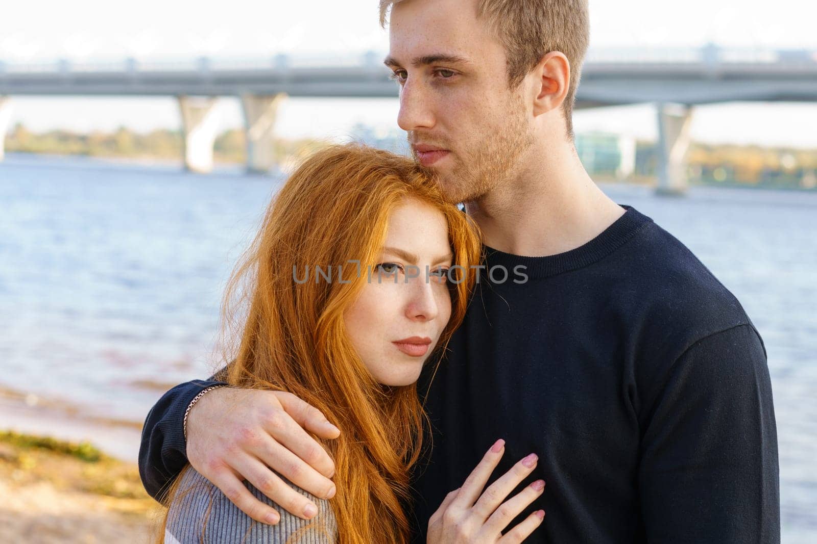 Young couple of man and woman with long red hair of Caucasian ethnicity, in casual clothes, stand on the bank of the river embracing happy on a summer day against the background of the cityscape