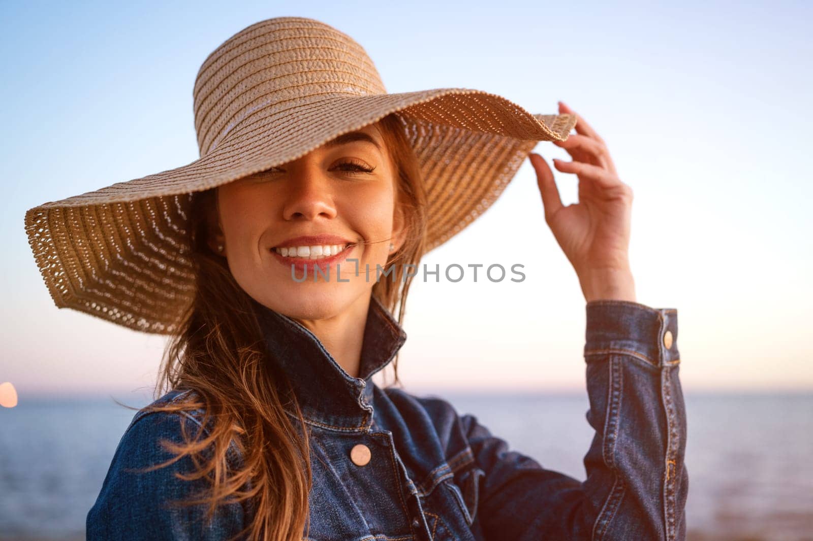 Portrait of a happy elegant young woman in a white denim jacket and a straw hat on the ocean shore at sunset, looking into the distance. Young caucasian female model close up on the seashore.
