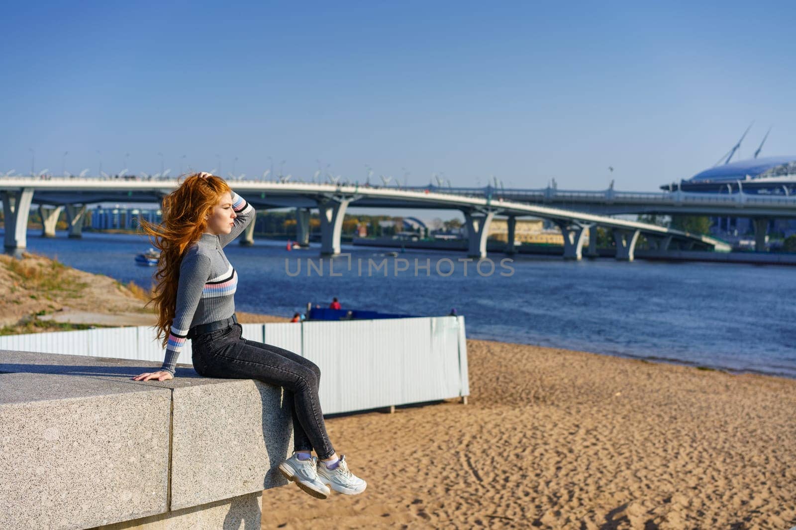 Beautiful redhead young woman sits on the embankment on a spring day by EkaterinaPereslavtseva