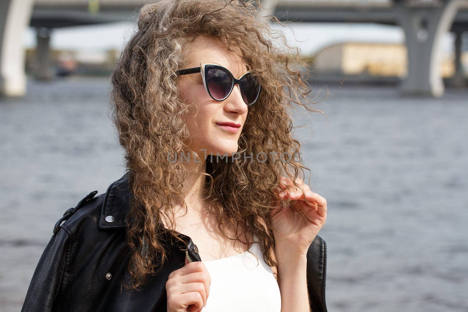 female portrait close-up, caucasian curly woman in a black leather jacket in sunglasses stands in the wind on the shore