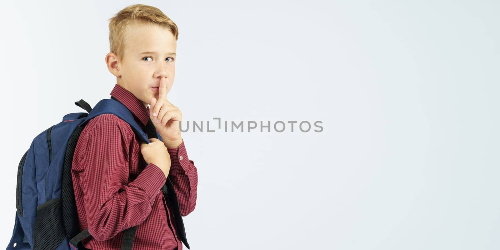 A schoolboy holds a school backpack and shows a gesture with his hand - be quiet. Education concept