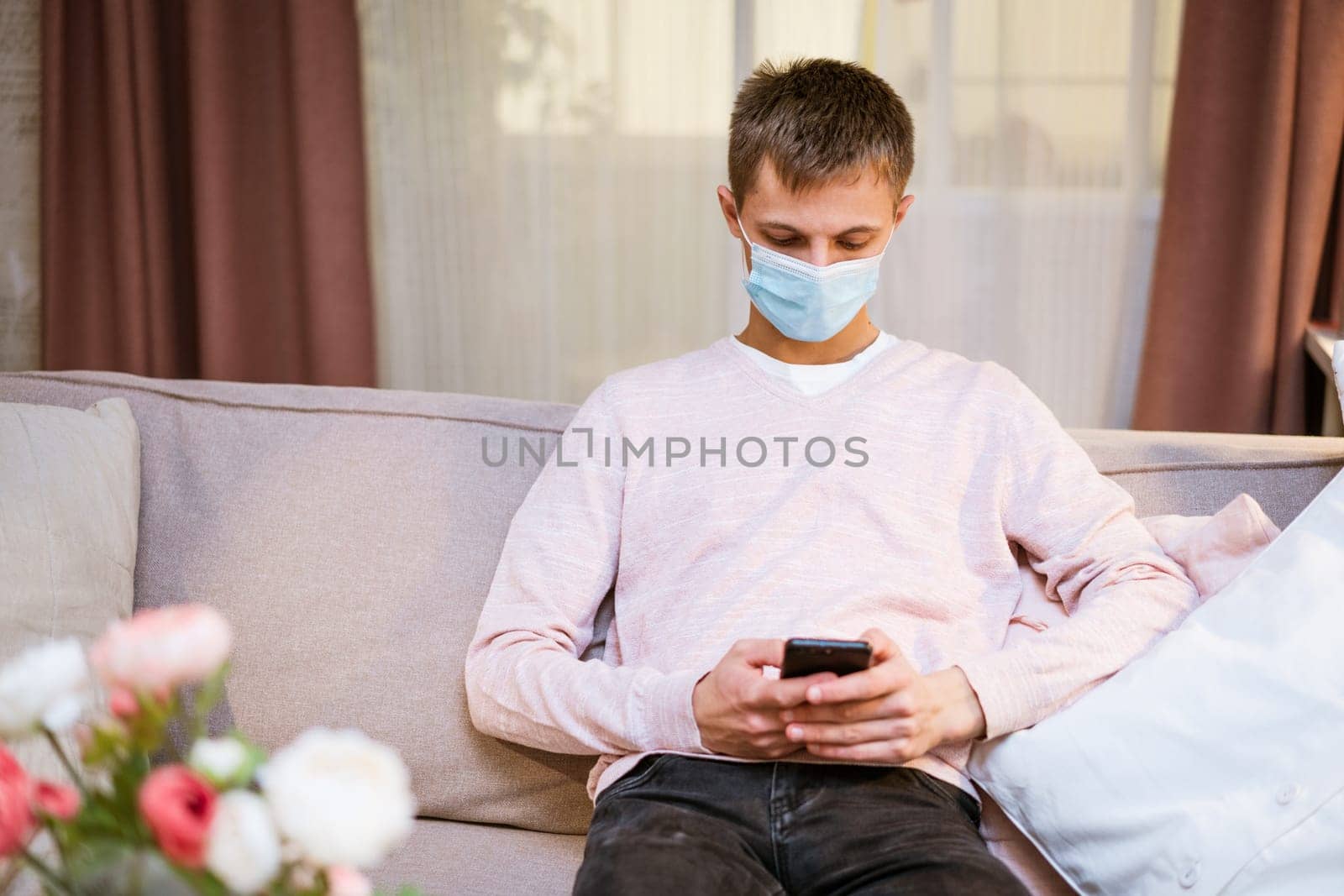 a young man sits on a sofa wearing a protective mask with a phone in his hand by EkaterinaPereslavtseva