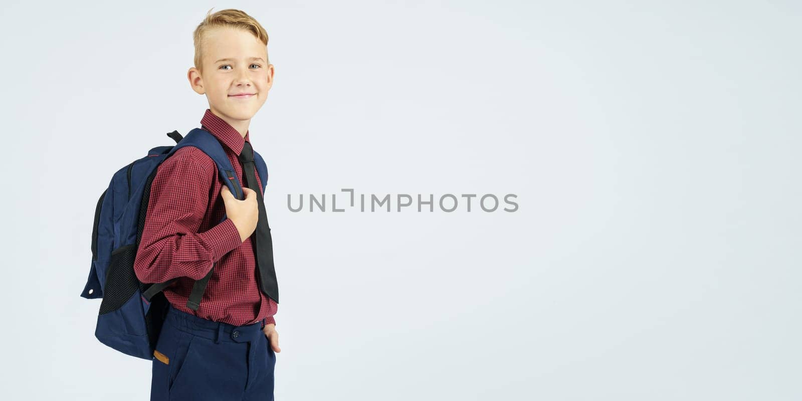 A schoolboy holds a school backpack, stands sideways, looks at the camera. Education concept