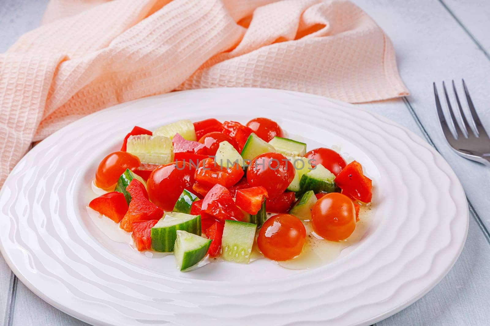 Salad of cucumbers, cherry tomatoes, peppers and dressing in a plate on a white wooden background