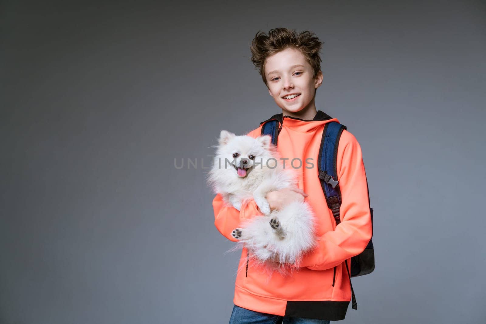 cheerful cute schoolboy of Caucasian ethnicity in bright clothes with a backpack and a dog in his hands on a gray background posing.