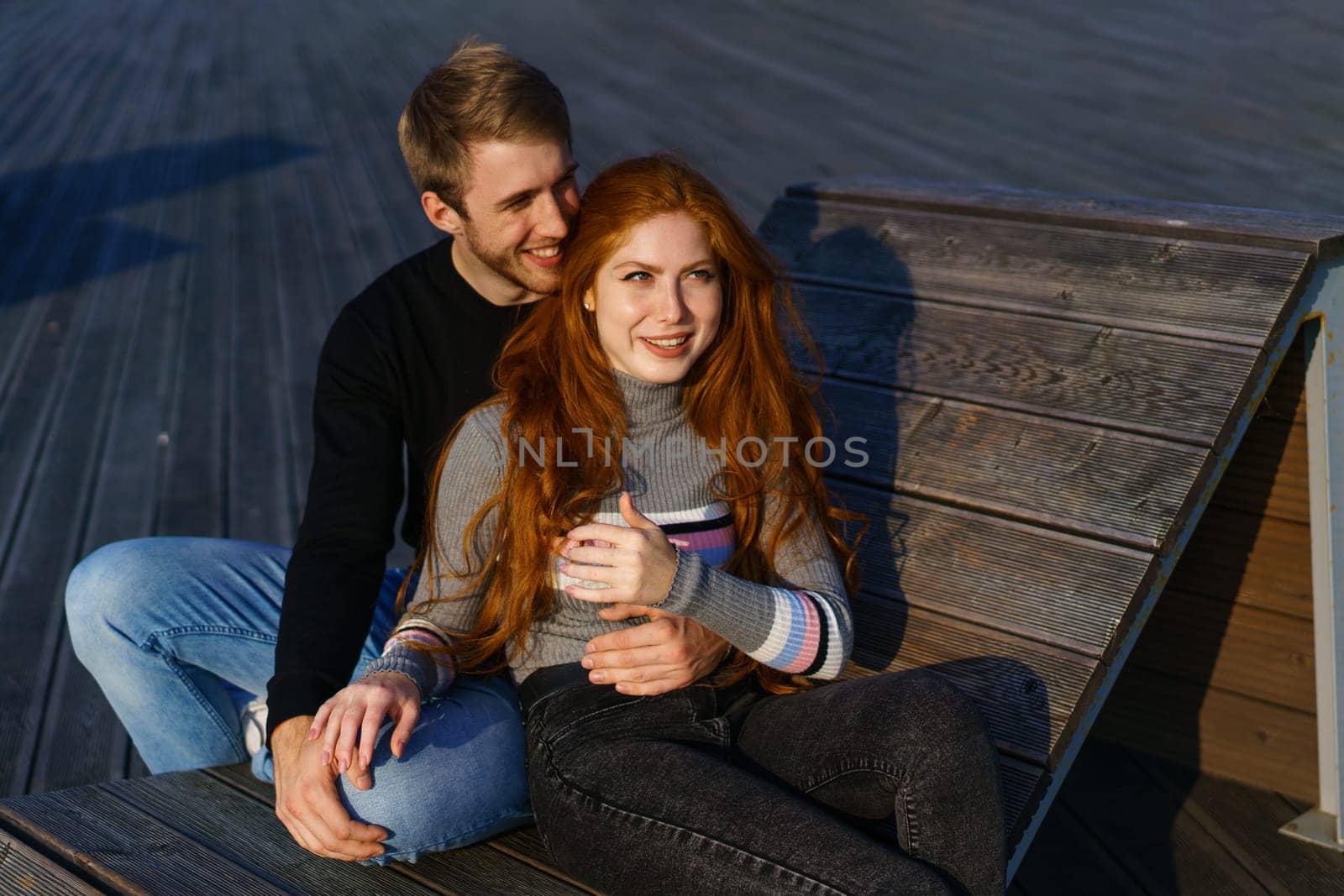 young couple of guy and girl with red hair of Caucasian appearance, in casual clothes, on a sunny day sitting in the park on a wooden bench in an embrace, happy relationship between a man and a woman