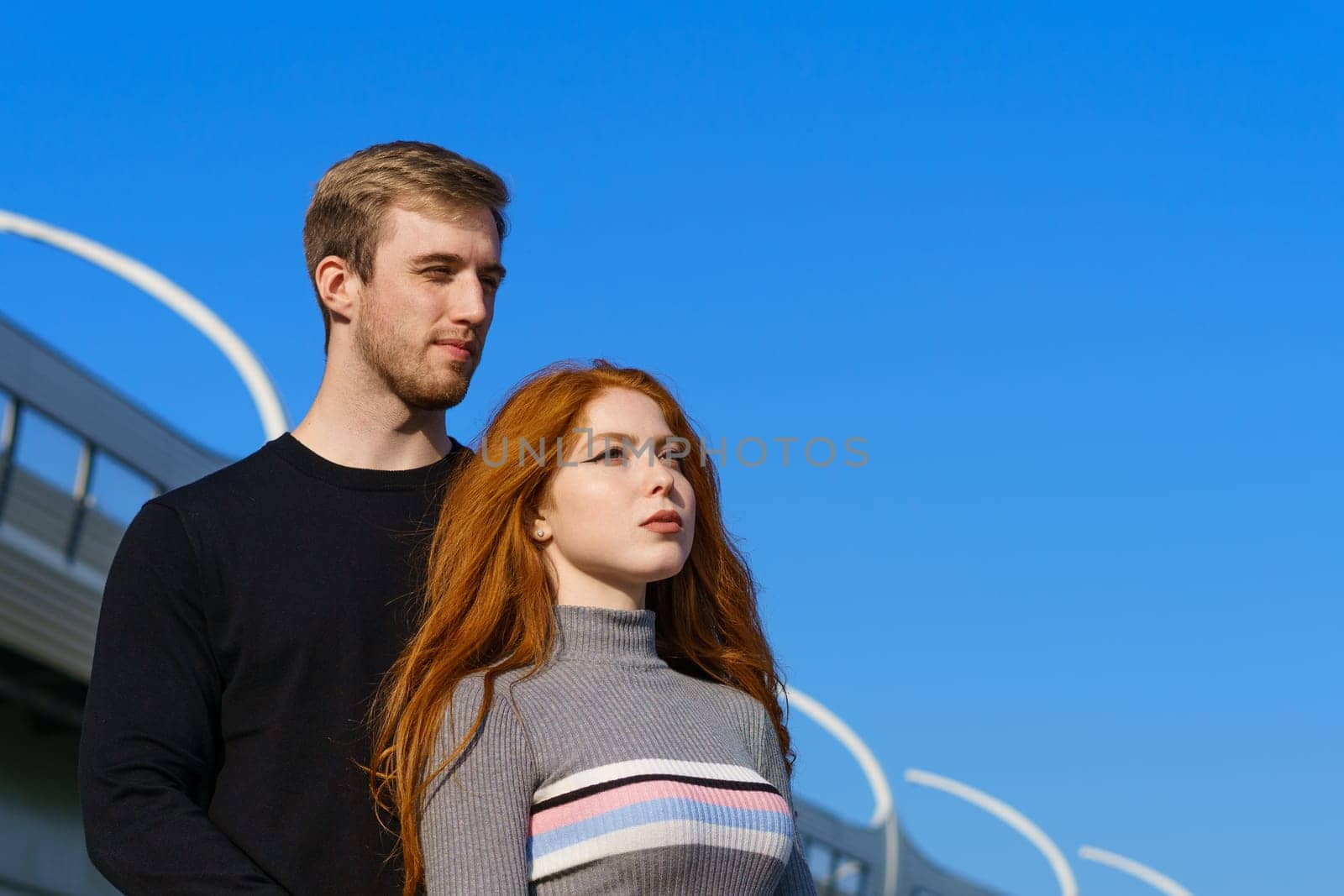 happy young couple man and woman with long red hair, stand against the background of a blue sky and a bridge in casual clothes and smile. Cheerful guy and Caucasian girl on a sunny day