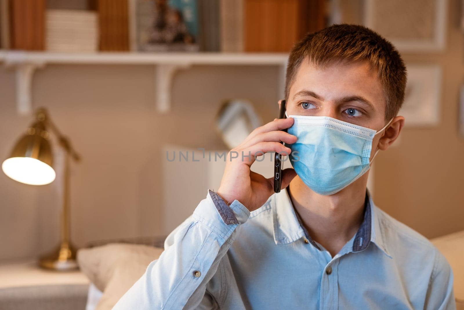 young man talking phone wearing protective mask while sitting on couch at home by EkaterinaPereslavtseva