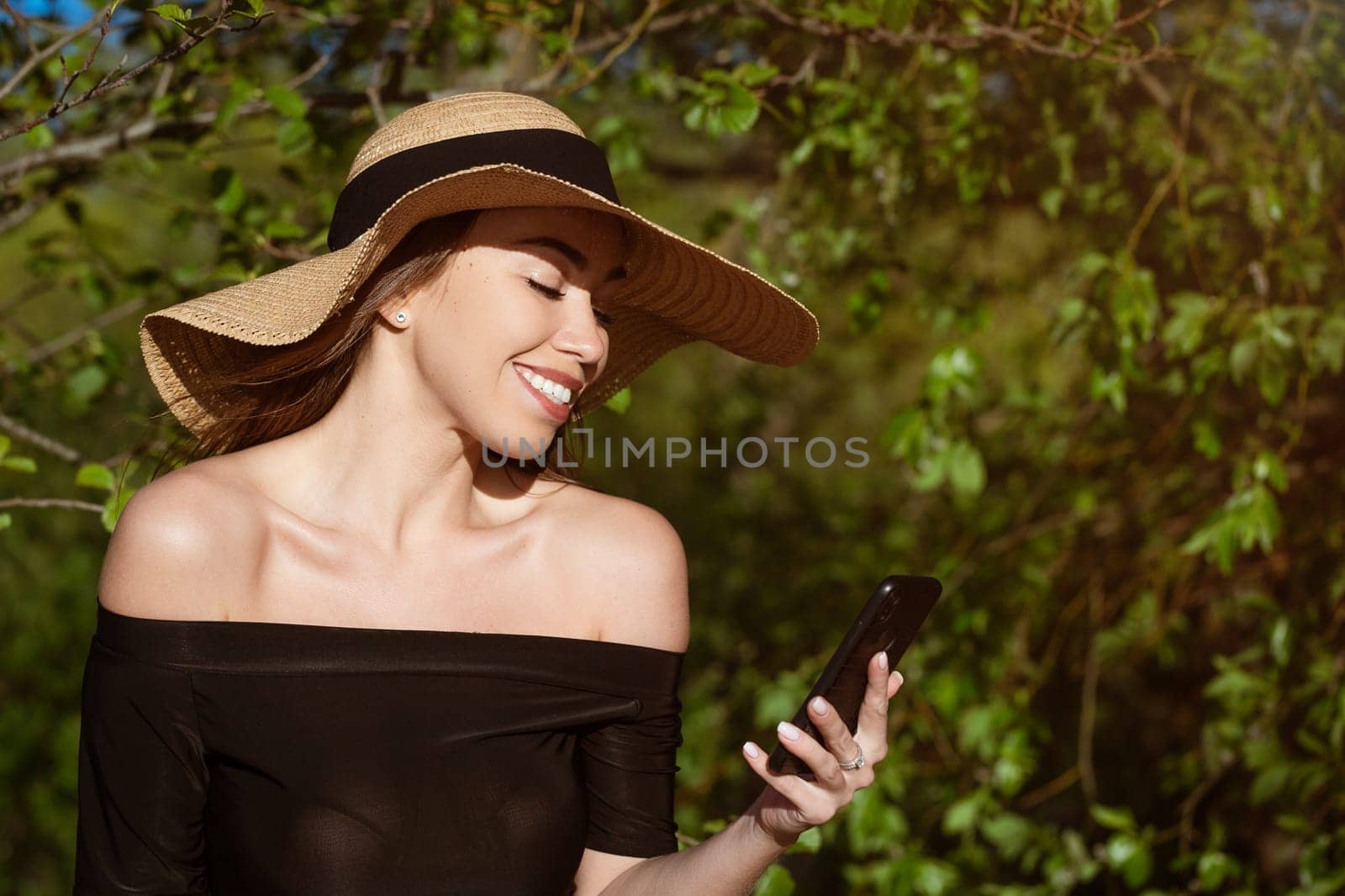 Beautiful happy young woman of caucasian ethnicity in a straw hat from the sun with large brim in a black dress with a phone in her hand smiling on a sunny summer day