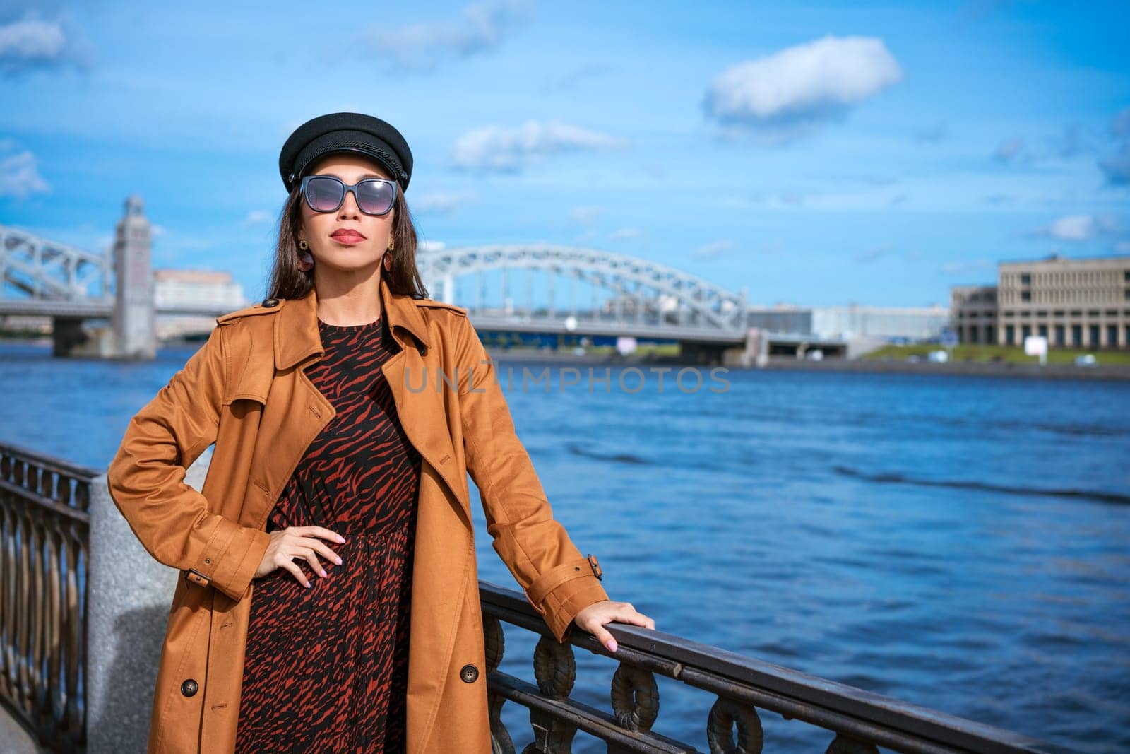 A young European woman in sunglasses and a black cap poses on the embankment in a brown coat on a spring sunny day. The concept of a successful and free woman