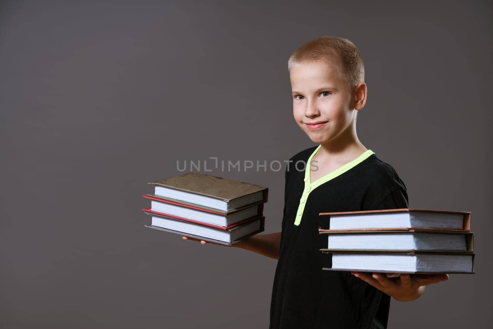 Cute boy in a black t-shirt holds stacks of books on a gray background
