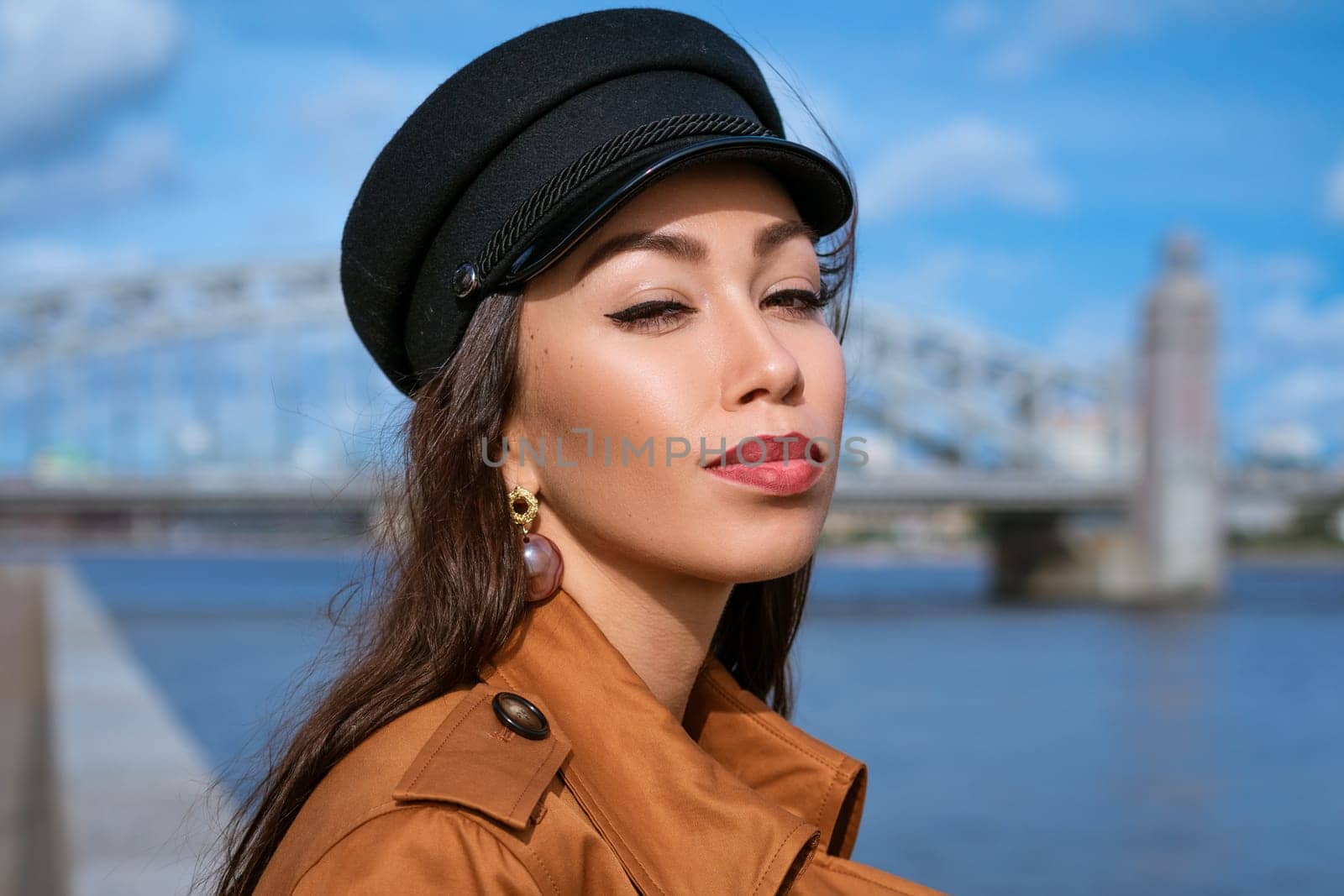 portrait of a happy Caucasian woman on the embankment of the river in a black cap and brown jacket on a sunny day