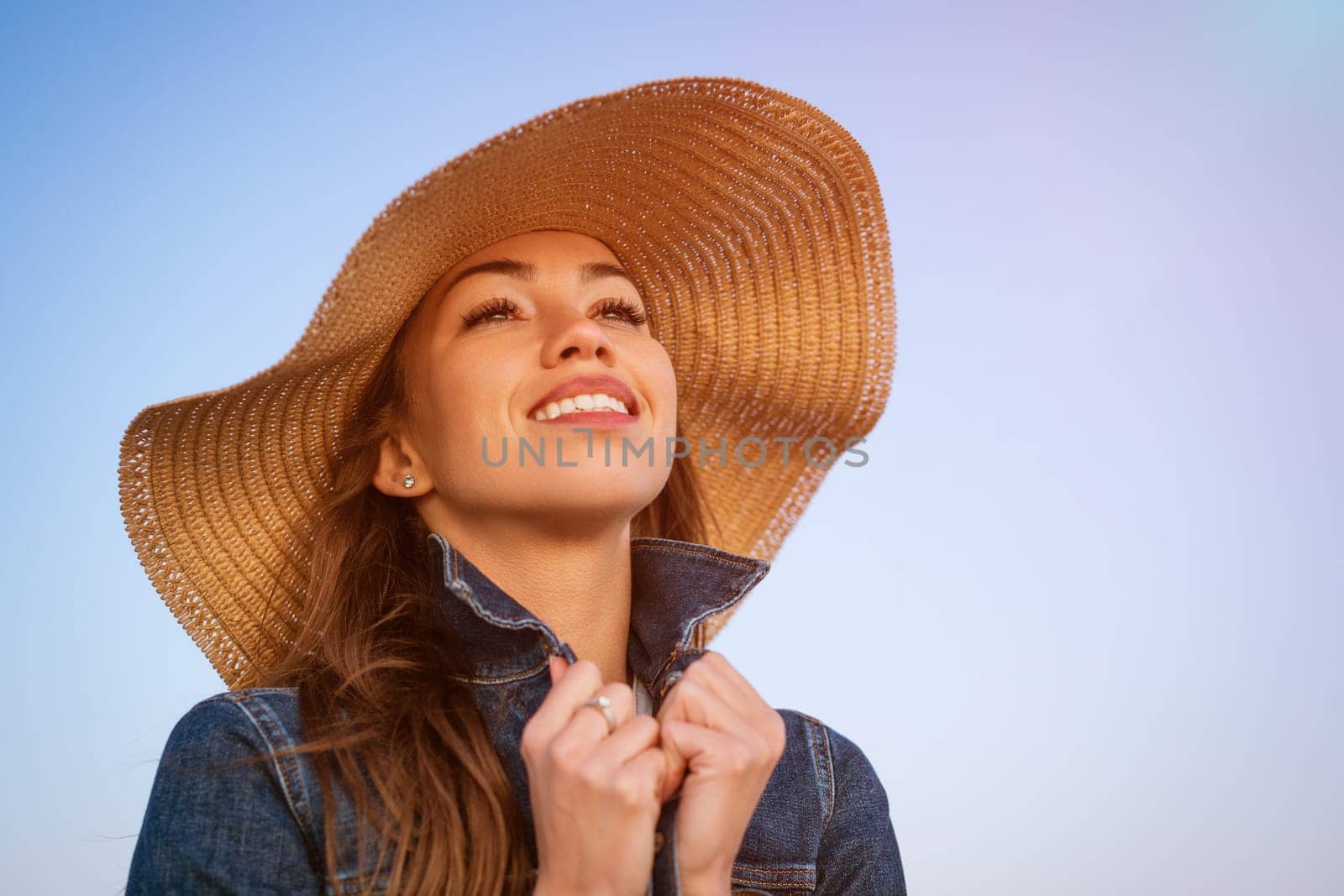 Portrait of a happy elegant young woman in a white denim jacket and a straw hat on the ocean shore at sunset, looking into the distance. young caucasian female model close up on the seashore.