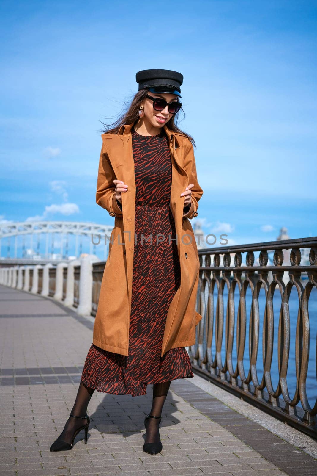 Beautiful caucasian woman in sunglasses, in a black cap and jacket posing while standing on the embankment on a sunny day against a background of blue sky and cityscape