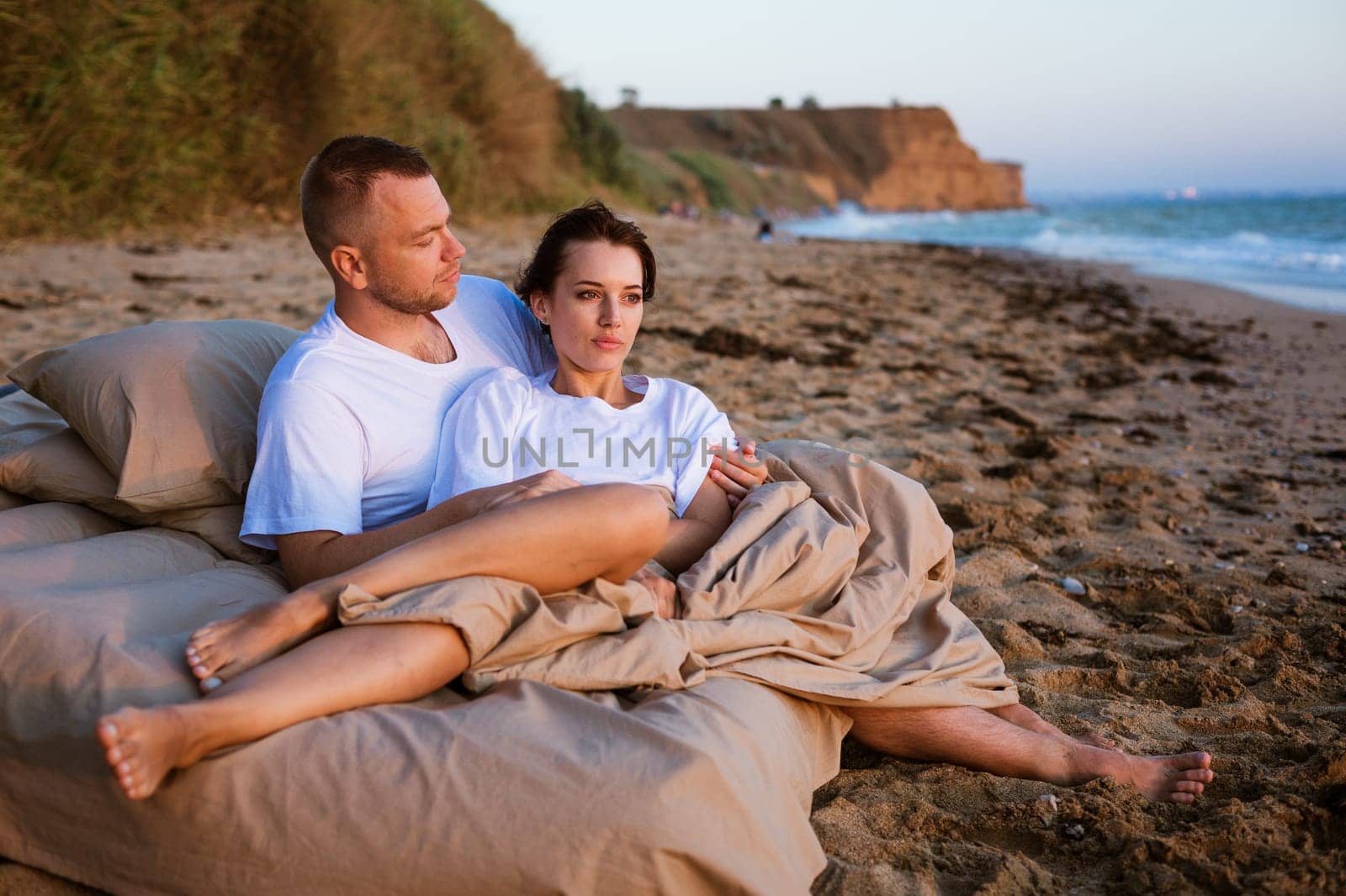 Young couple wrapped in a blanket outdoors. Happy couple in love lying and rotating in bed by the sea at sunset. Happy caucasian couple of husband and wife wrapped in a blanket outdoors