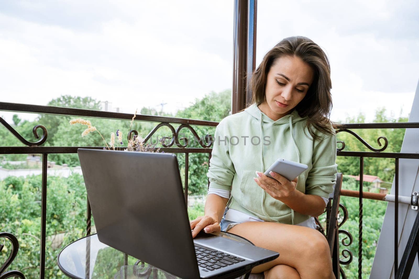 Young woman sits on a balcony at a table working with a phone behind a laptop by EkaterinaPereslavtseva