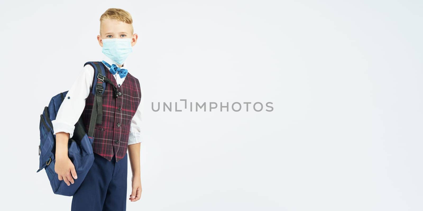 A schoolboy with a medical mask on his face holds a school backpack, stands sideways, looks at the camera. Education concept