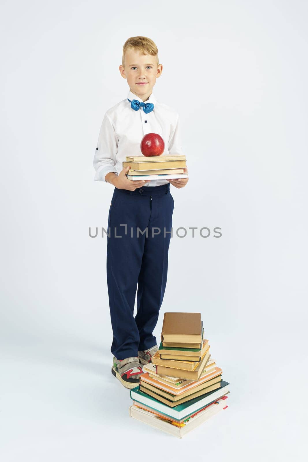 A schoolboy stands near a stack of books, holding books and an apple in his hands. Isolated background. Education concept