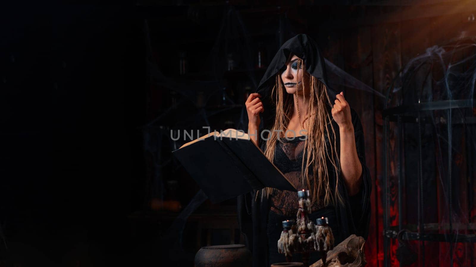 Halloween concept. Witch dressed black hood standing dark dungeon room use flying magic book for conjuring magic spell. Female necromancer wizard gothic interior making curse