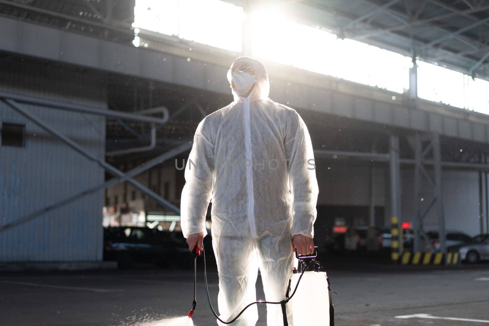 Man dressed white protective overalls spraying surface antibacterial sanitizer sprayer during quarantine Caucasian person protective suit disinfects parking during covid pandemic backlight