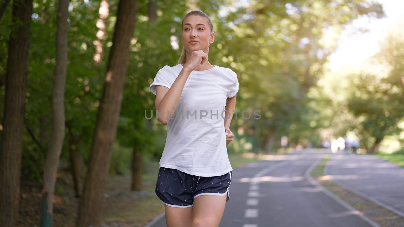 Woman running asphalt road summer park Active sporty caucasian female morning workout Healthy lifestyle concept. Athletic person sportswear exercises Dressed white t-shirt