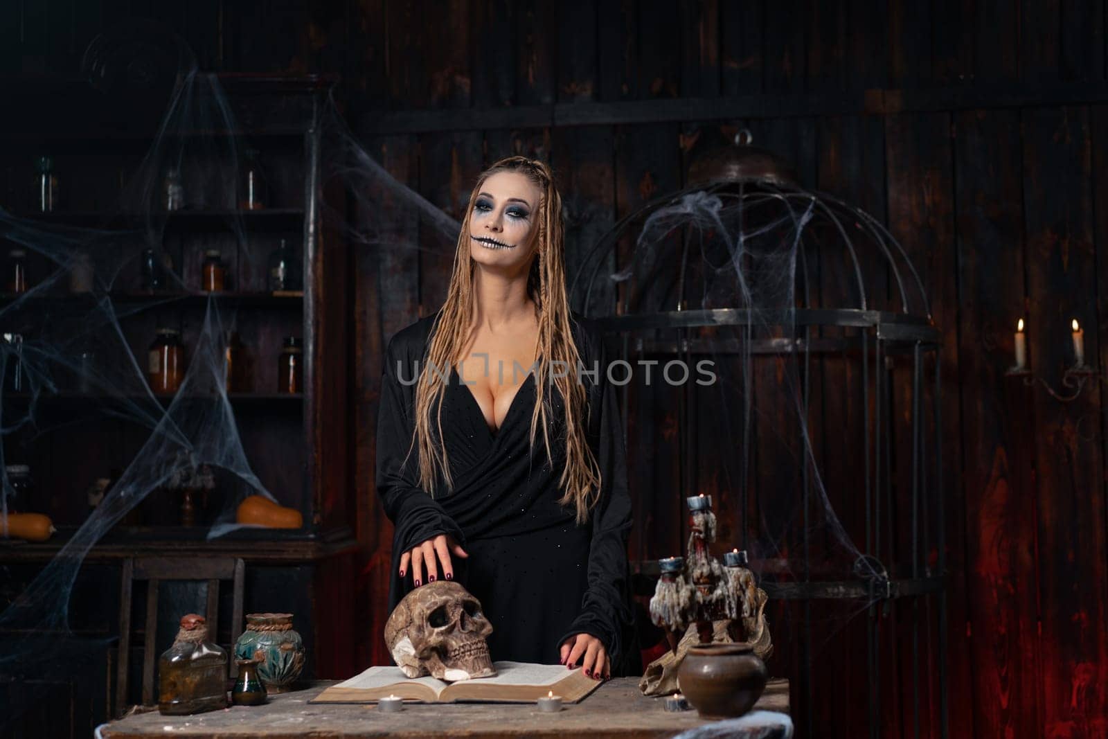 Halloween concept. Witch portrait close up with dreadlocks holding human scull in hand dressed black hood standing dark room occultism religion Caucasian female necromancer