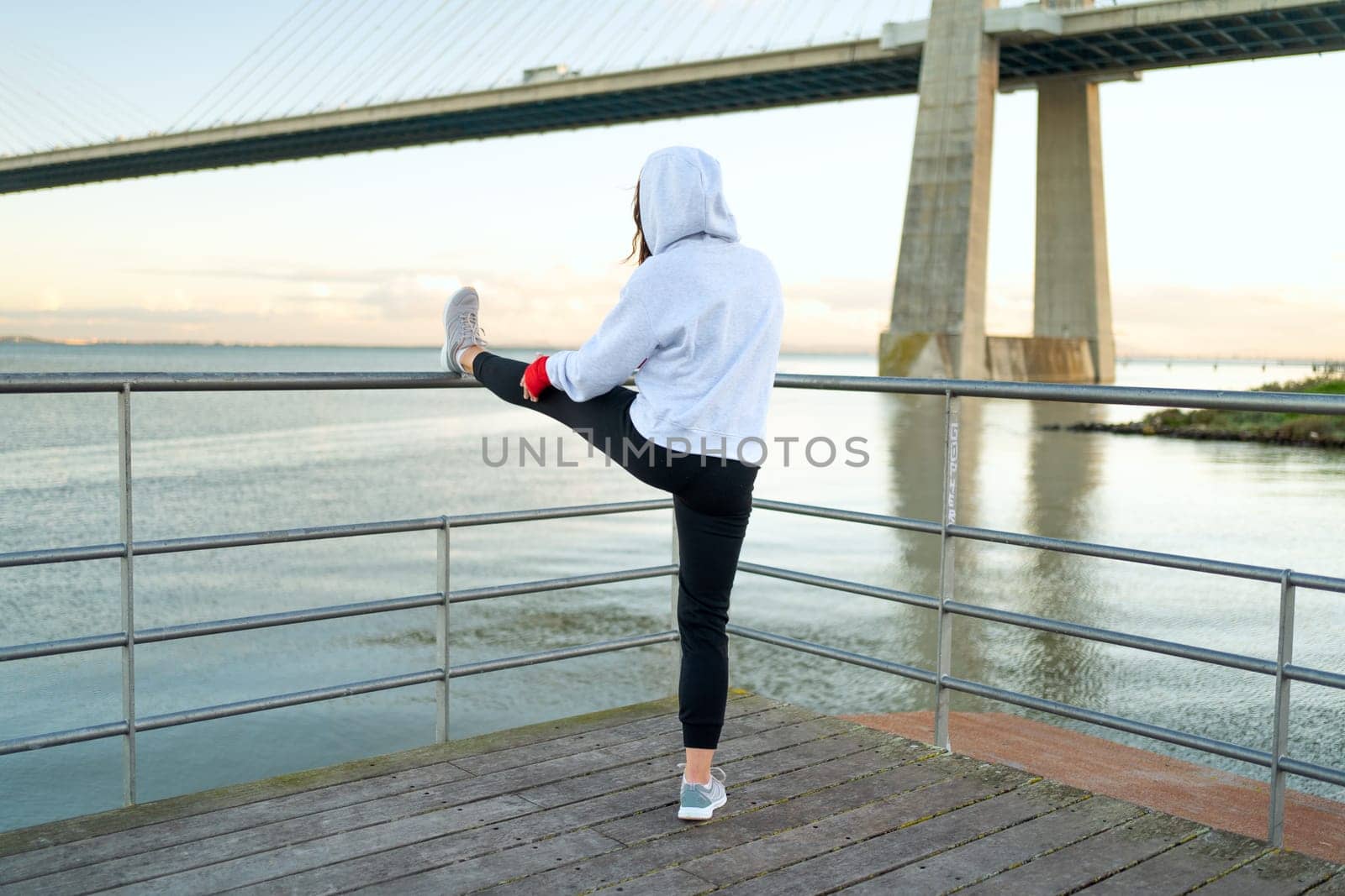 Athlete age woman stretching legs before kickboxing exercises. Caucasian athletic female does lunges exercises stretch leg seaside view with bridge and blue sky background. Back view