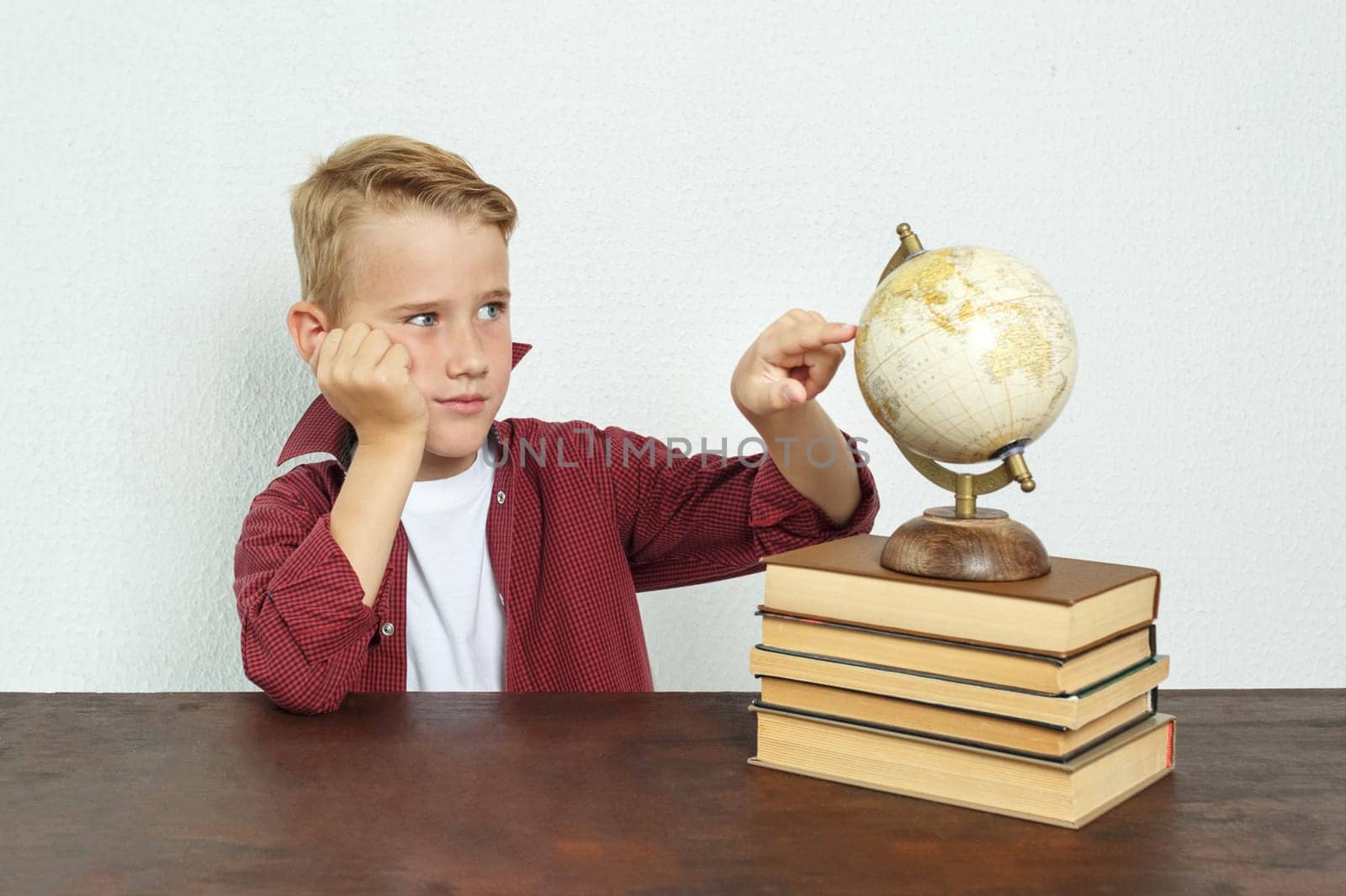 Education concept. The schoolboy sits at the table, propping his head on his hand, points to the globe. On the table there are books, a globe and an alarm clock.