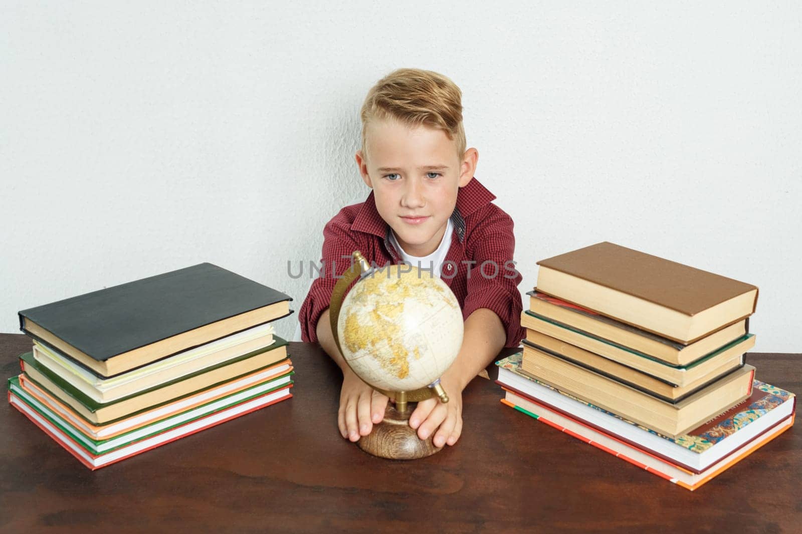 Education concept. The schoolboy sits at the table, shows the globe. On the table there are books, a globe and an alarm clock.