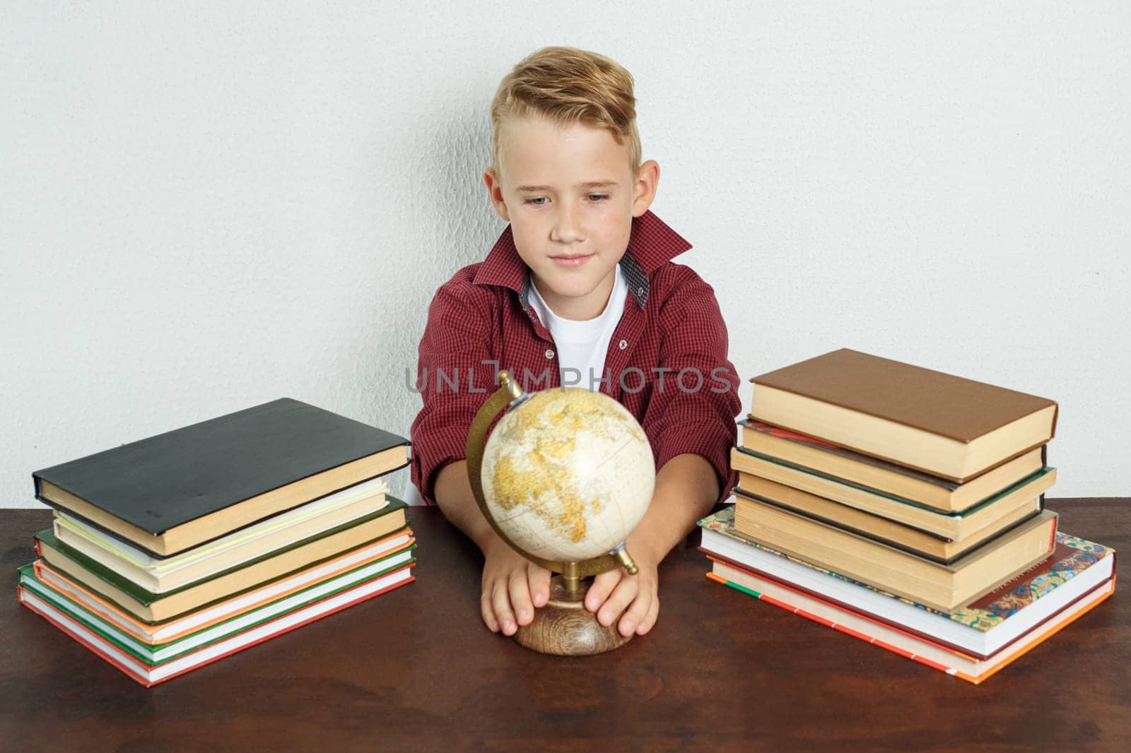 Education concept. The student sits at the table and looks at the globe holding on to it with his hands. On the table there are books, a globe and an alarm clock.