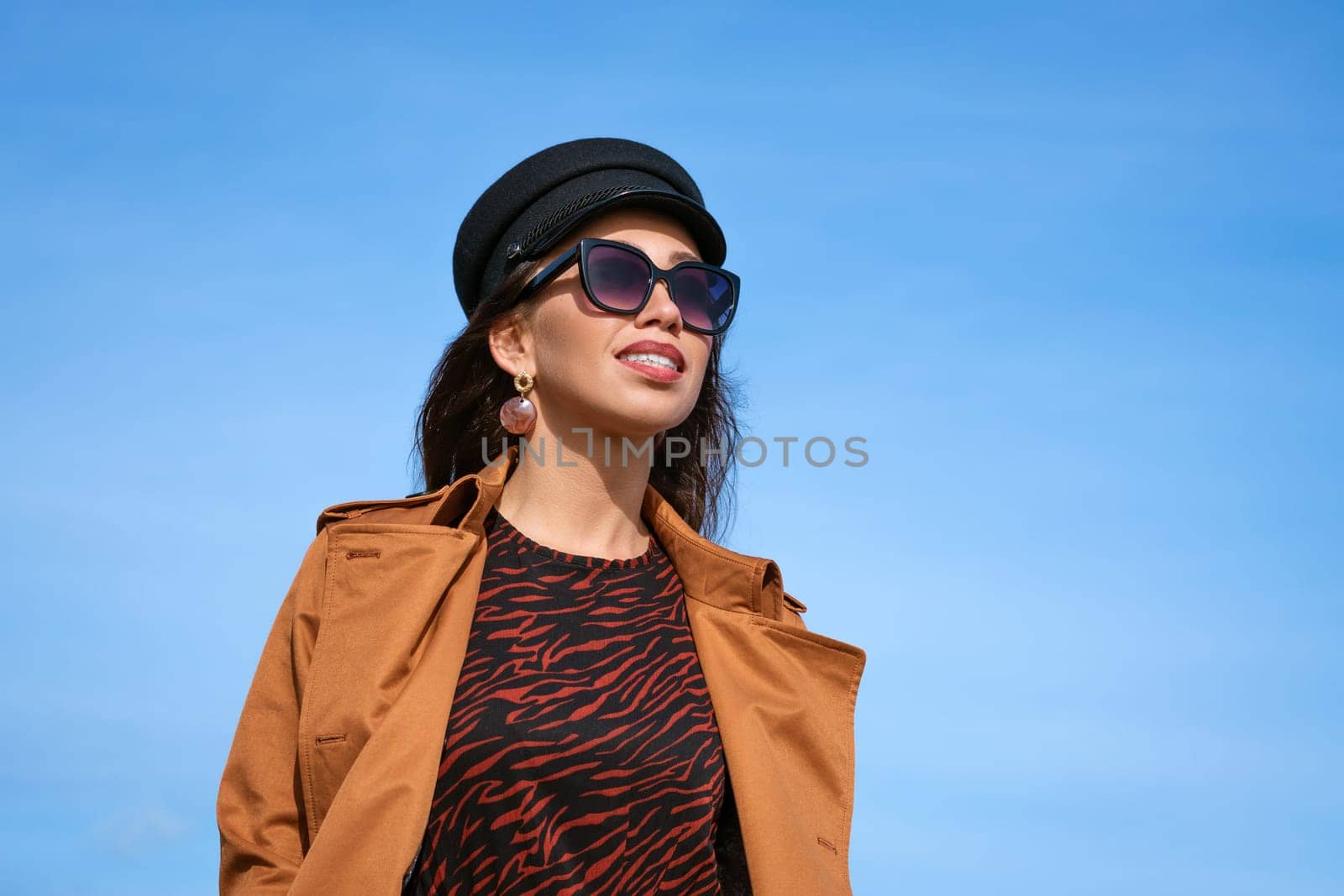 Female portrait of a beautiful young woman of Caucasian appearance in a black cap against a blue sky in sunglasses and a jacket