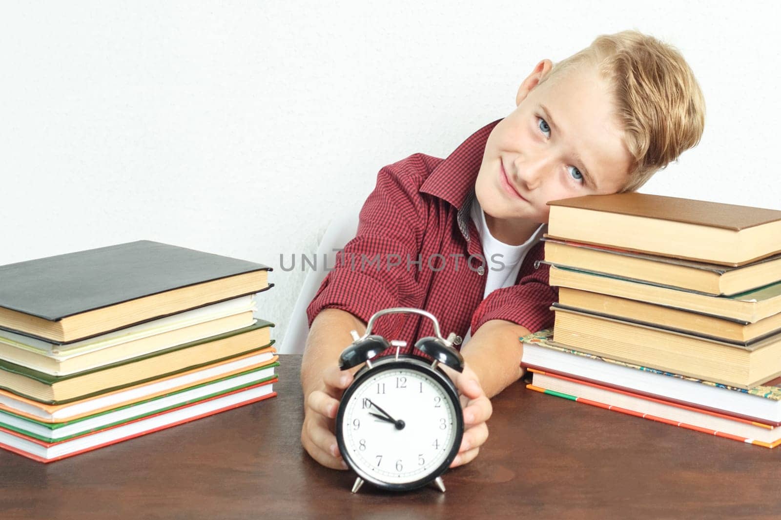 Education concept. The schoolboy sits at the table and holds an alarm clock in his hands.