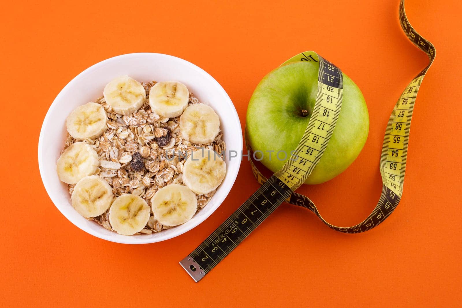 muesli with banana in a white plate on an orange background, green apple with a yellow measuring tape. Healthy food and diet concept