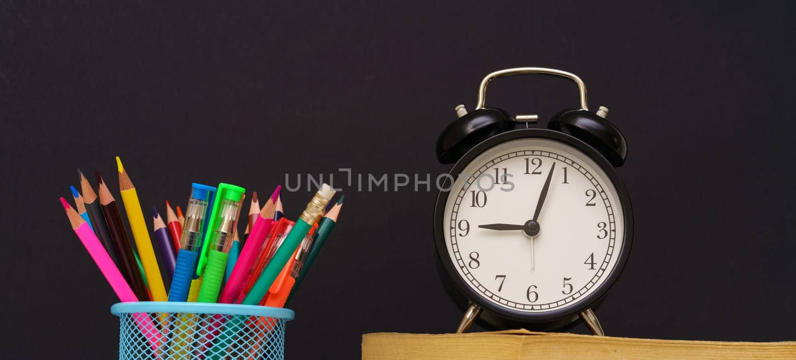 the alarm clock stands on the books and glass with pencils on black background, concept back to school