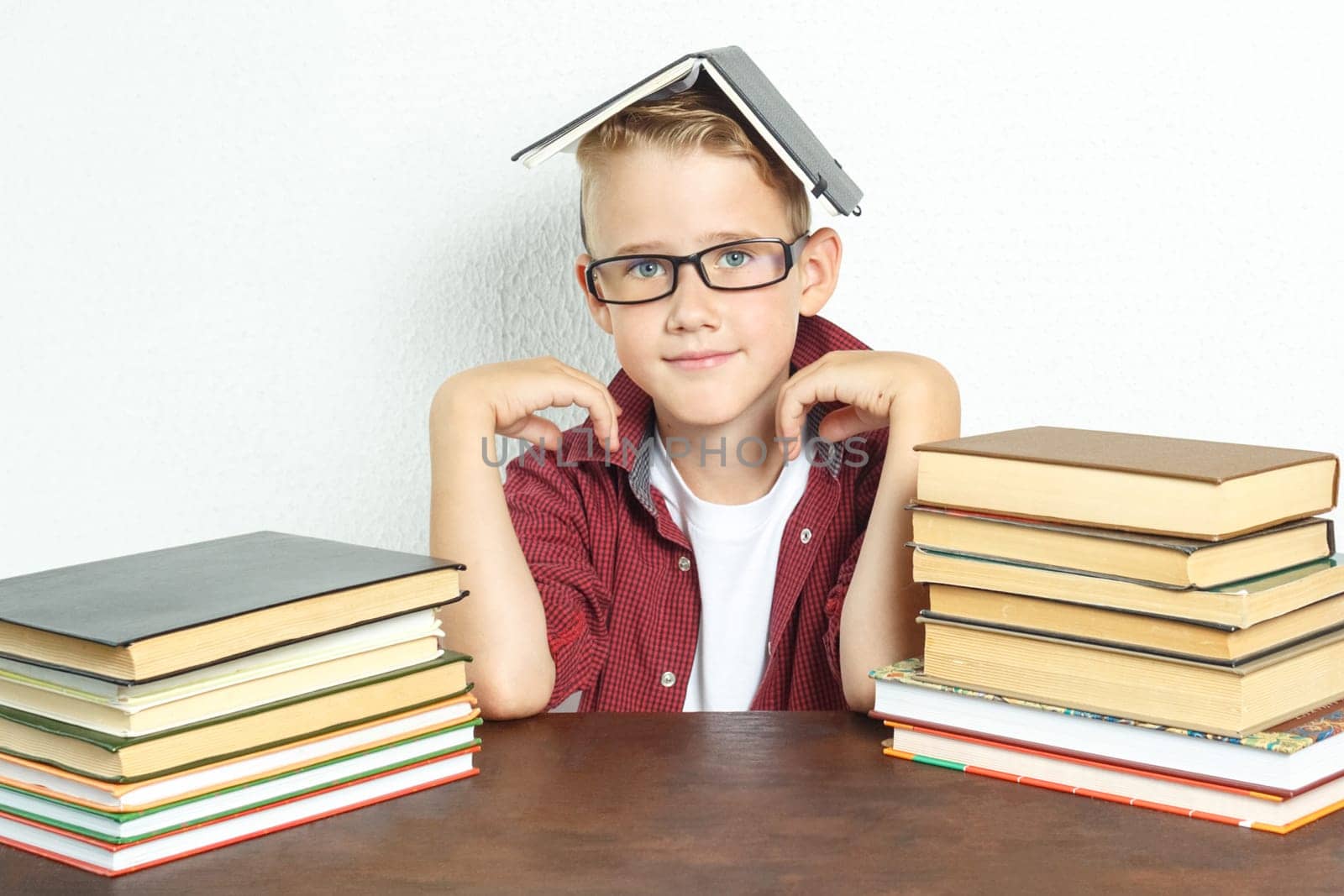 Education concept. A pupil boy sits at a table with books, holding an open book on his head.