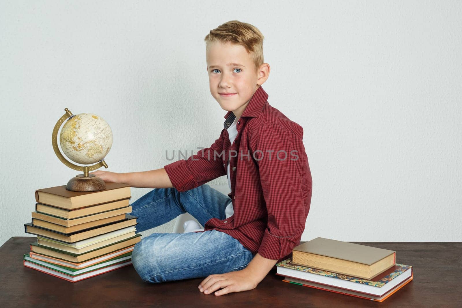 A schoolboy sits at a desk near books and holds a globe with his hand. Education concept