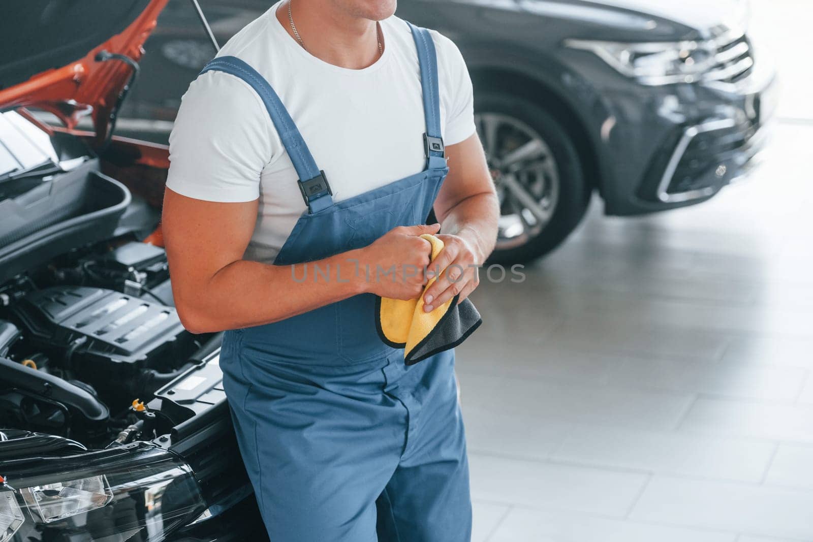 In blue uniform. Man is repairing broken automobile indoors.
