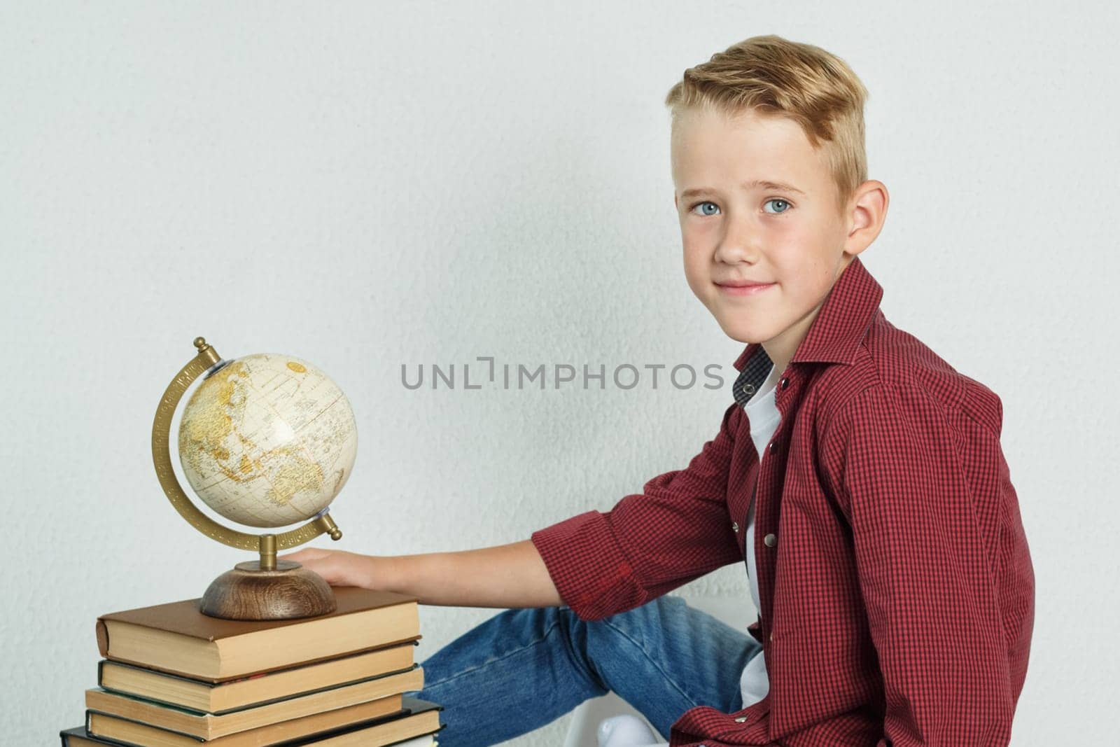 A schoolboy sits at a desk near books and holds a globe with his hand. Education concept