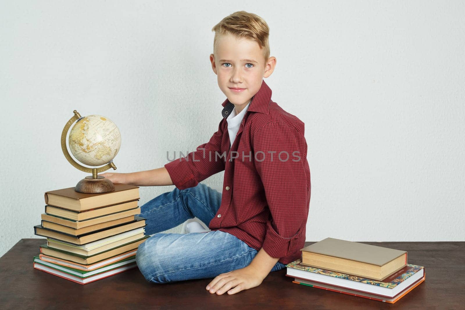 A schoolboy sits at a desk near books and holds a globe with his hand. Education concept