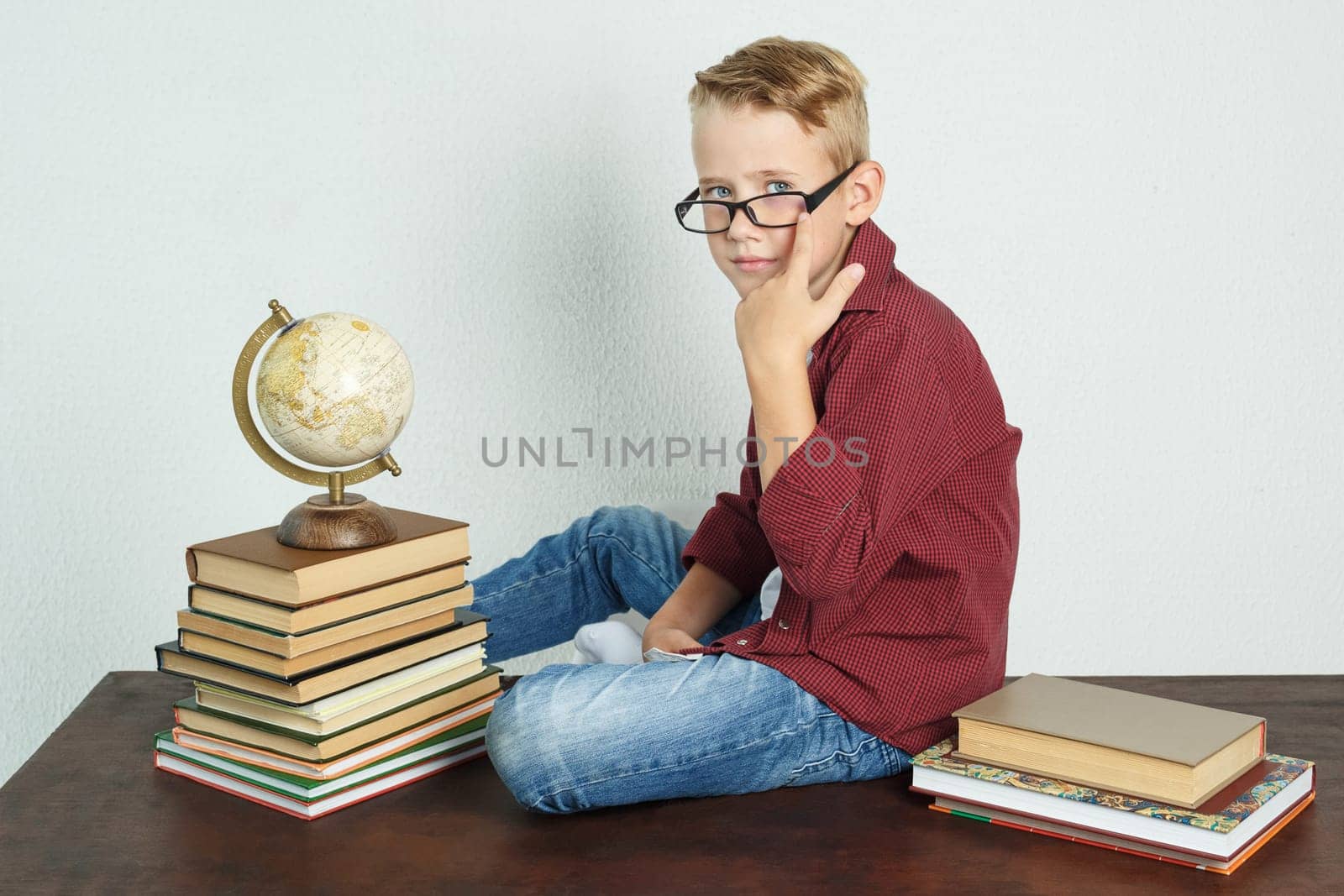 A schoolboy sits on a table near books and a globe, straightens his glasses. Education concept