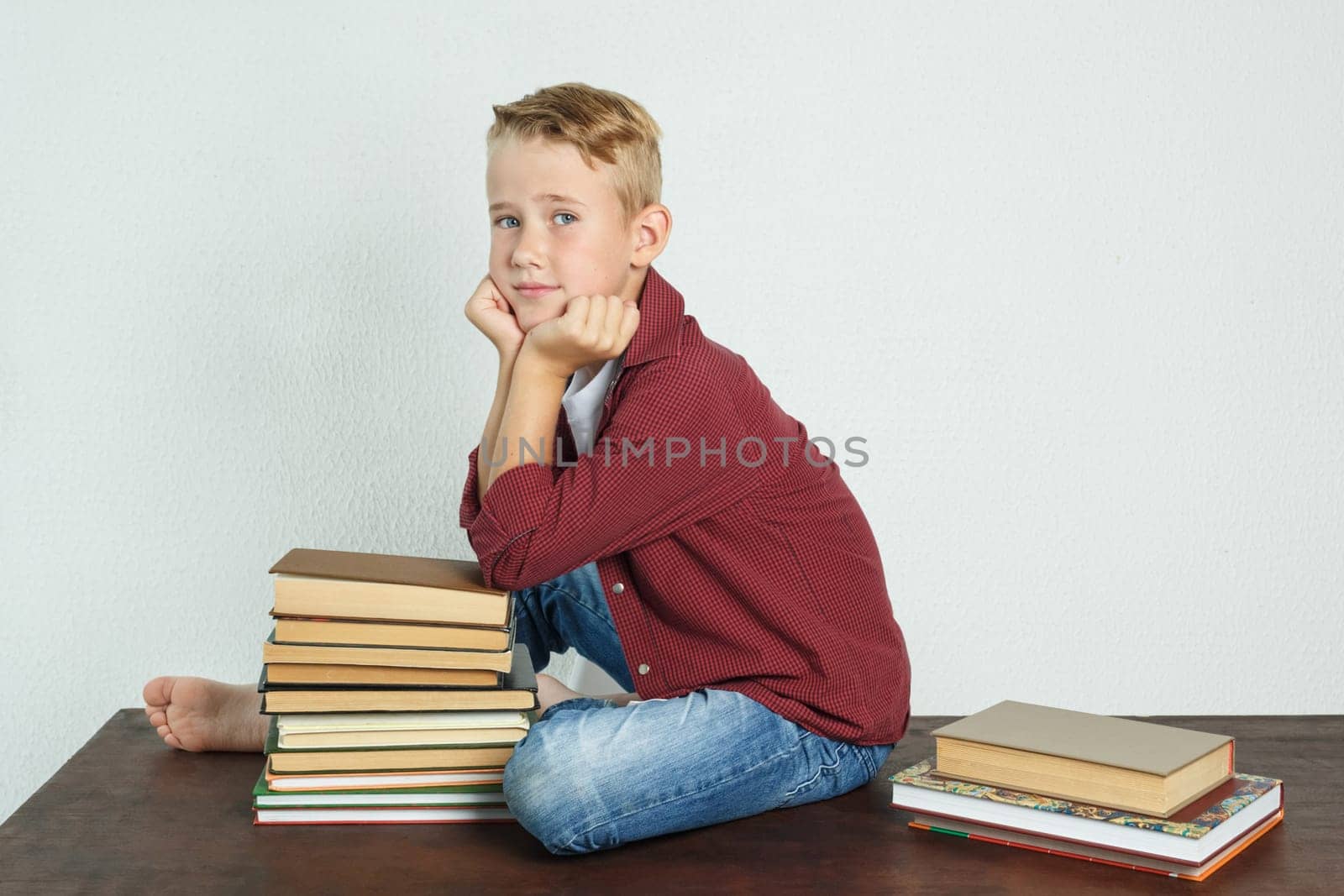 A schoolboy sits on the table near the books, leaning his elbows on them and looks into the cameras. Education concept