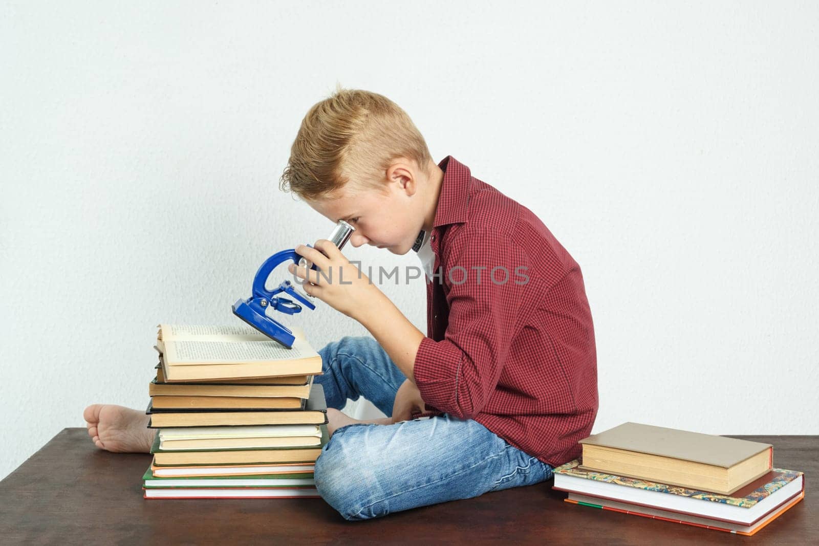 A schoolboy sits at a table near books and looks through a microscope. Education concept