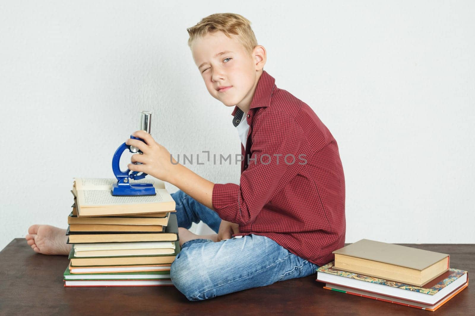 A schoolboy sits at a table near books, holds a microscope in his hands and looks at the camera. Education concept