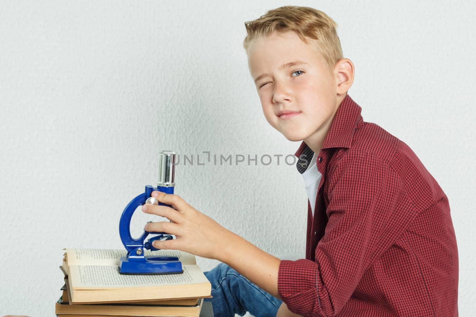 A schoolboy sits at a table near books, holds a microscope in his hands and looks at the camera. Education concept