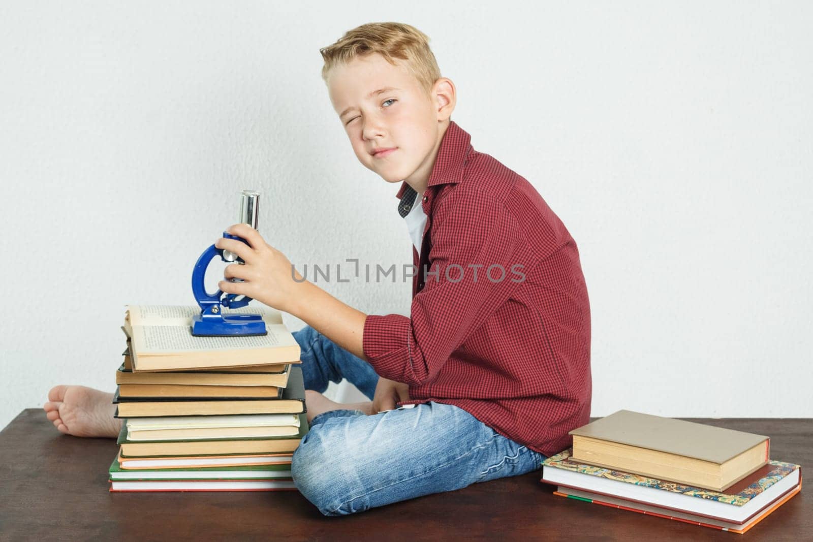 A schoolboy sits at a table near books, holds a microscope in his hands and looks at the camera. by Sd28DimoN_1976
