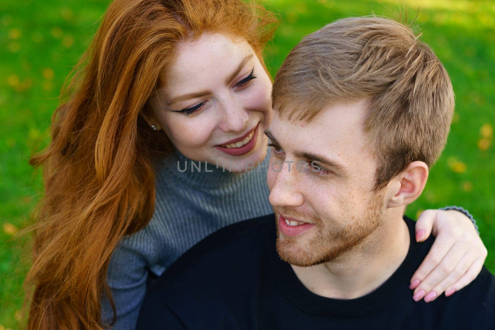 Happy young couple of Caucasian ethnicity man and woman in casual clothes sitting embracing in the park on green grass on a sunny day. Happy relationship between prana and a girl in nature