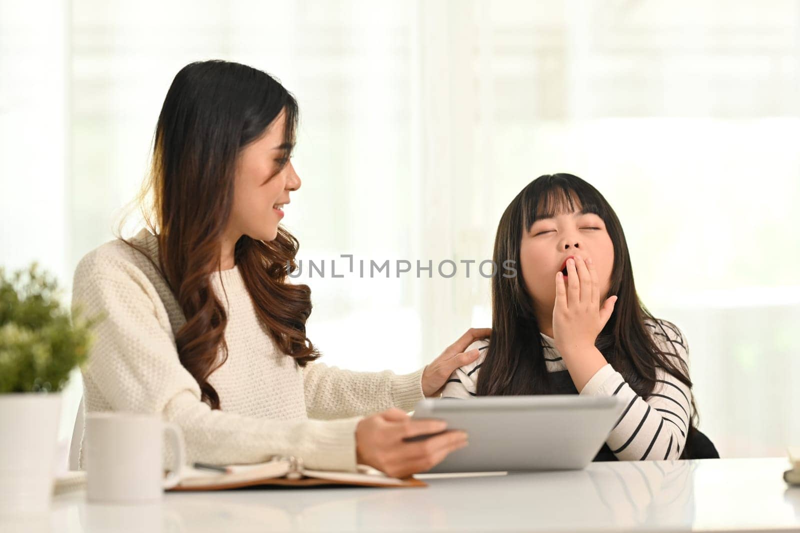 Shot of sleepy little girl yawning, covering mouth with hand during doing homework at home with her mother.
