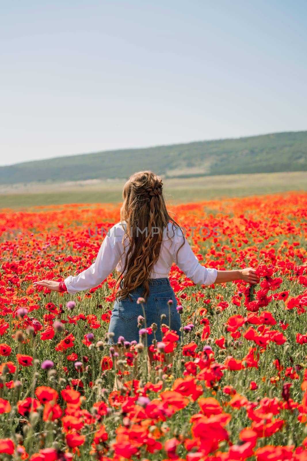 Happy woman in a poppy field in a white shirt and denim skirt with a wreath of poppies in her hand posing and enjoying the poppy field. by Matiunina