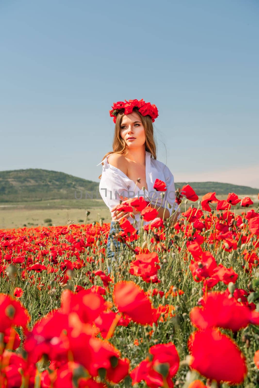 Happy woman in a poppy field in a white shirt and denim skirt with a wreath of poppies on her head posing and enjoying the poppy field