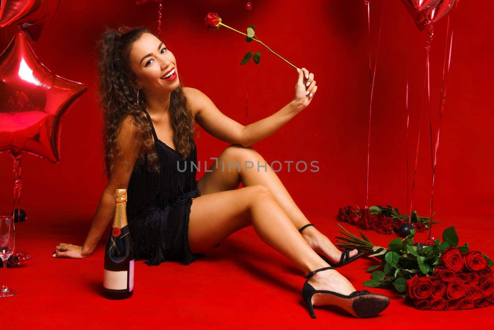 Girl posing sitting on a red background with roses and a bottle of champagne. A young woman of Caucasian ethnicity in a black dress sits on a background with red balloons and cute smiles